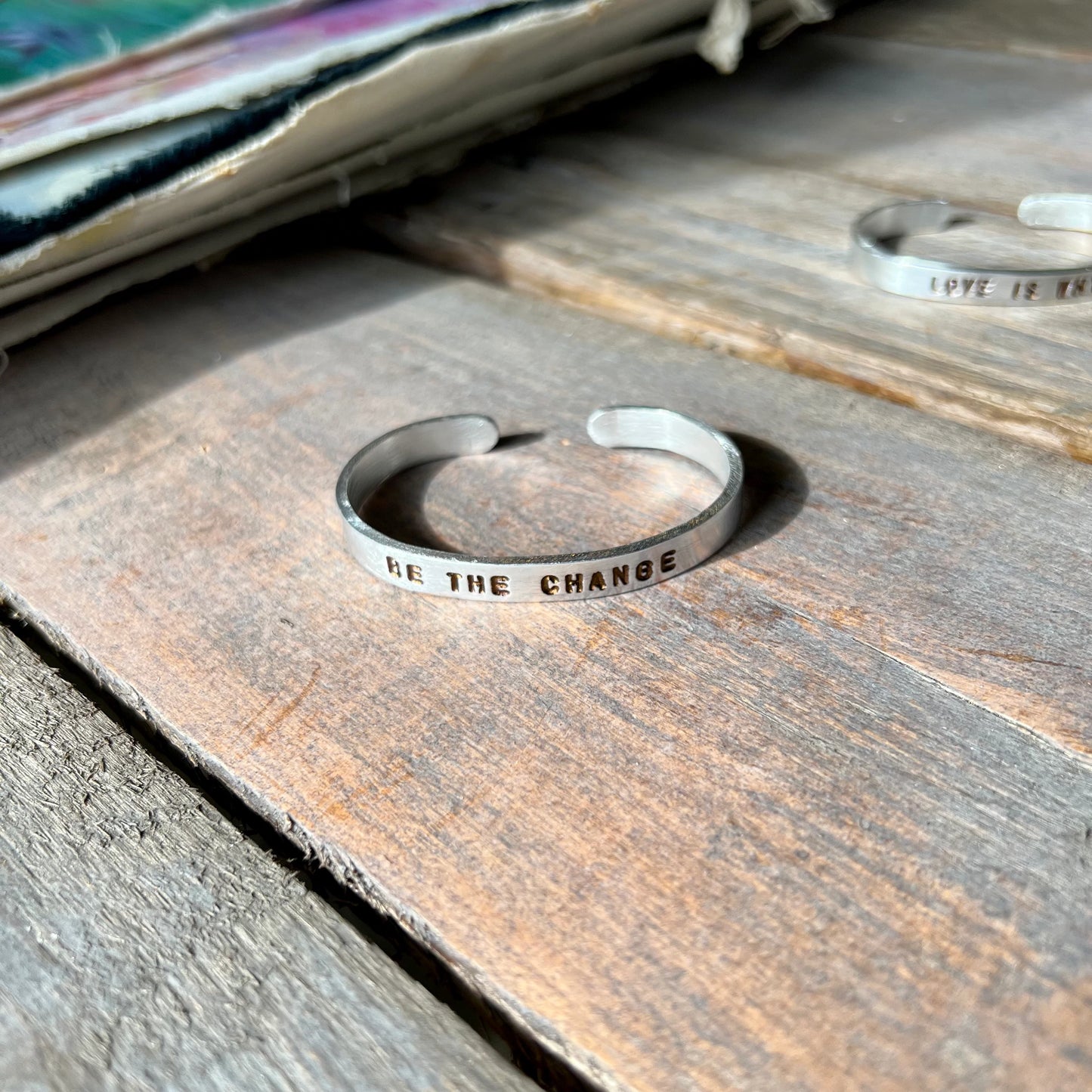 Silver Be The Change Cuff with stamped and hammered text on an adjustable aluminum band